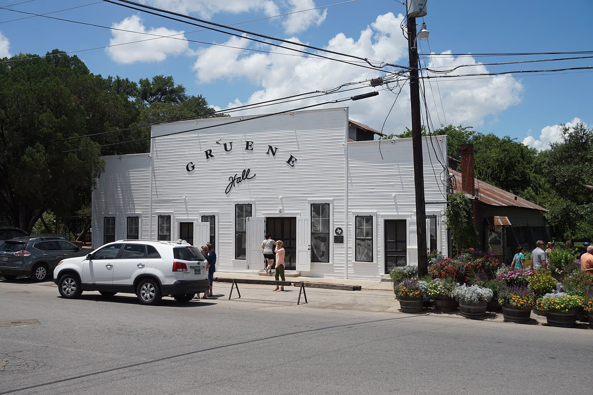 The white clapboard exterior of Gruene Hall with its painted false-front sign, the Texas historical marker plaque, and whiskey-barrel flower planters out front under a blue sky